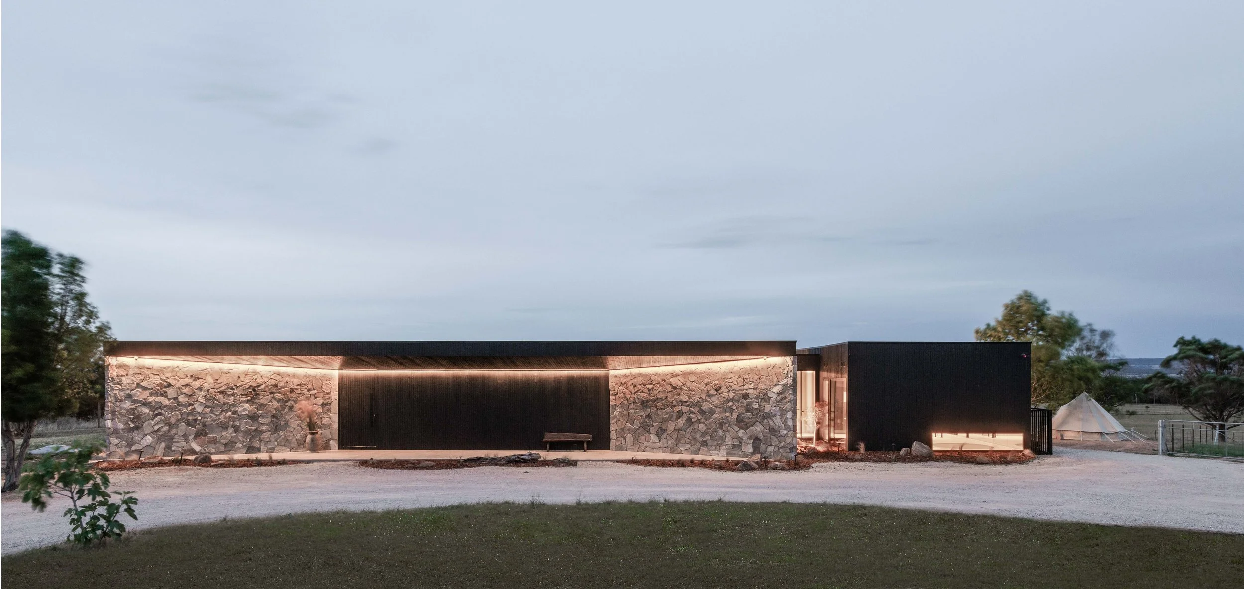 Zensi Retreat — front exterior, stone wall and blackened-timber pavilion at dusk